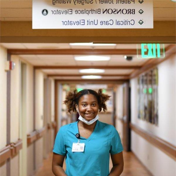 Mariah Rudolph stands in hospital hallway in blue scrubs and face mask pulled down to chin