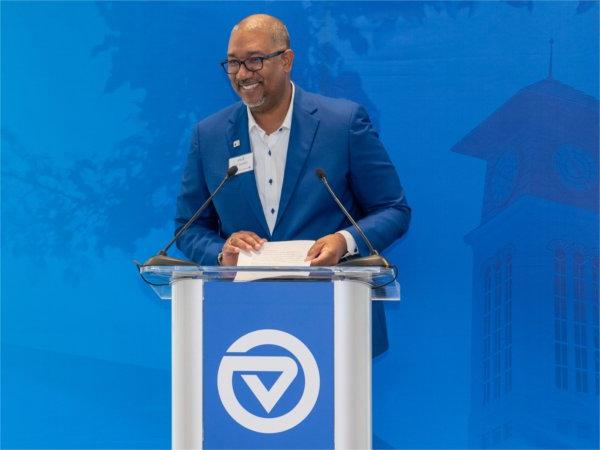 A man in a blue suit stands at a GVSU podium and smiles as he addresses the crowd.